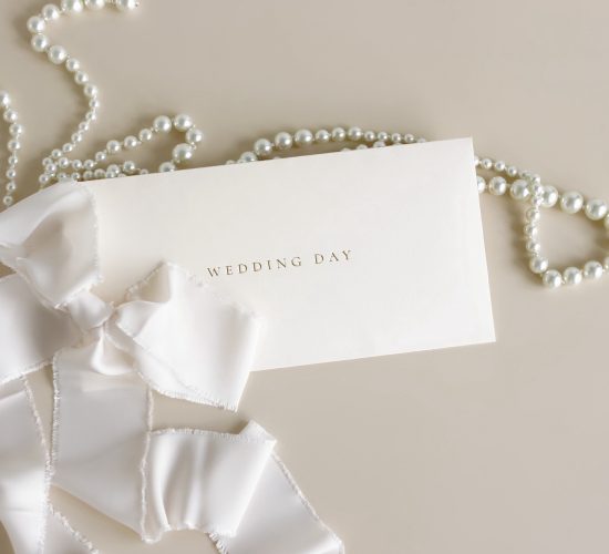 Wedding invitation envelope, greeting card mockup with white silk ribbon and pearl necklace on beige background top view.Copy space. Wedding backdrop.